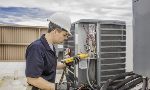 Trained hvac technician holding a voltage meter, performing preventative maintenance on a air conditioning condenser unit.